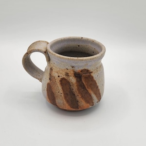May include: Handmade ceramic mug with a rounded body and a sturdy handle. The mug features a speckled, light-colored exterior with brown, diagonal stripe designs. The interior is a light purple color. A unique piece for enjoying coffee or tea.