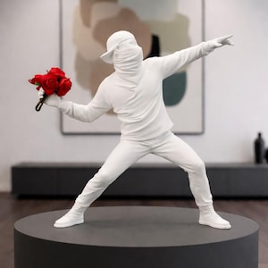 Könnte beinhalten: Eine weiße Skulptur einer Figur mit Baseballkappe und Gesichtsbedeckung, die einen Strauß roter Rosen in der einen Hand hält und die andere ausstreckt. Die Figur befindet sich in einer Wurfpose, auf einem dunkelgrauen kreisförmigen Sockel.