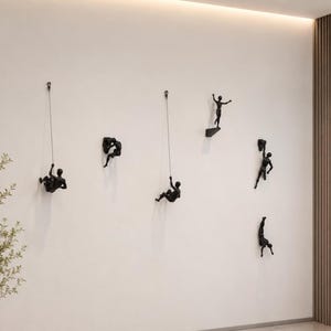 Könnte beinhalten: Ein Set aus sechs schwarzen Wandskulpturen, die Figuren in verschiedenen Kletter- und Sprungposen darstellen. Zwei Figuren hängen an dünnen Drähten, eine sitzt auf einem kleinen Vorsprung und drei sind direkt an der Wand befestigt.