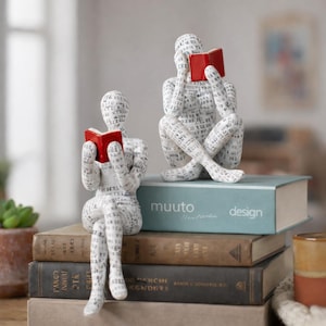 Peut inclure: Deux sculptures blanches de personnages lisant des livres à couvertures rouges. Les personnages sont recouverts de texte et sont assis sur une pile de livres. Les livres ont différentes couleurs et tailles, avec le mot "design" sur l'un d'eux.