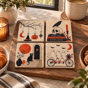 May include: A set of four square coasters featuring Halloween illustrations. One shows hanging charms, one a skull on books with the text Strange People, Spooky Places, Beautiful Seasons, one a black cat by a door, and one three ghosts on a bicycle.