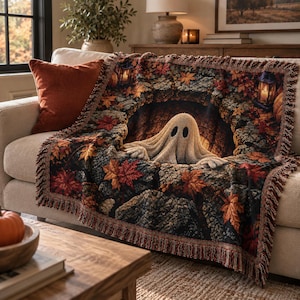 May include: A woven throw blanket featuring a central cream-colored ghost figure emerging from a dark, arched stone opening. The design includes vibrant autumn maple leaves in shades of red, orange, and yellow, with two glowing lanterns.