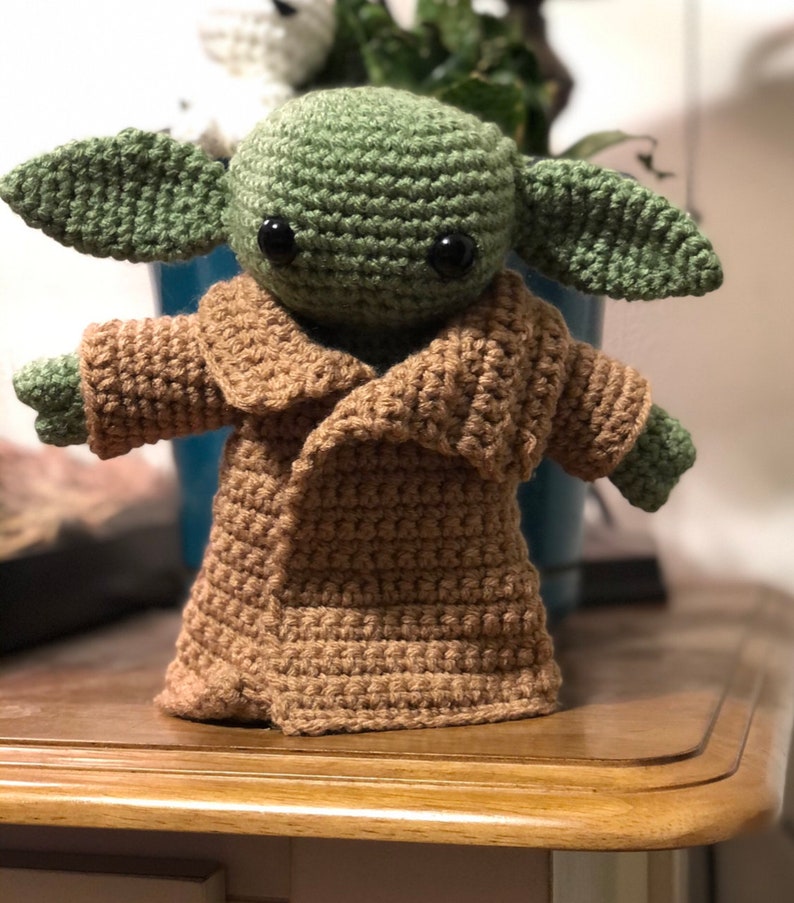 May include: A crocheted green and brown plush toy resembling a character from the Star Wars franchise. The toy has large ears, big eyes, and is wearing a brown coat.