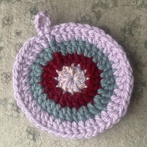 May include: A handmade, circular crochet potholder in shades of lavender, gray, burgundy, and white. The potholder features a loop for hanging and a concentric circle design. This item is suitable for kitchen use.