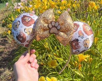 Op de afbeelding: Gouden glitter Minnie Mouse oren met een gouden strik en een pootprint bedel. De oren zijn versierd met een witte achtergrond en een patroon van Simba en andere personages uit The Lion King.