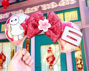 May include: Pink and white Minnie Mouse ears with a red sequin bow and a white Baymax character on one ear. The other ear has a white number 8 on it.