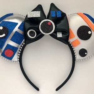 May include: A pair of black mouse ears with a black bow in the center. The ears are decorated with images of R2-D2 and BB-8 from Star Wars.