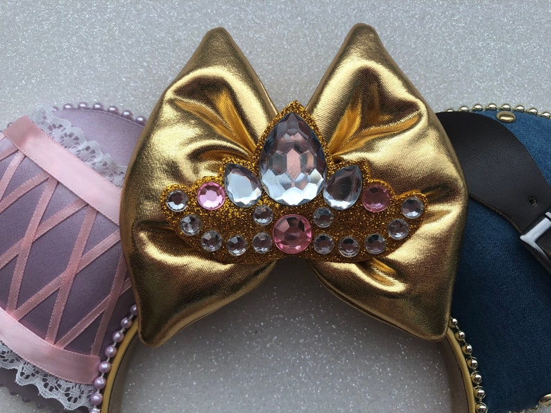 Puede incluir: Lazo dorado con un dise&ntilde;o de corona enjoyada, con diamantes de imitaci&oacute;n transparentes y rosas. El lazo est&aacute; unido a una diadema con una oreja de tela rosa y blanca a la izquierda y una oreja de mezclilla azul a la derecha.