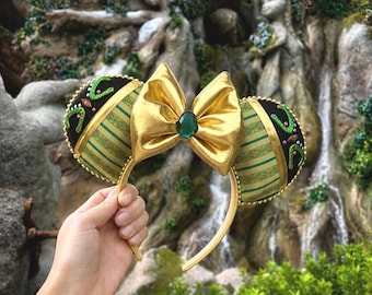 May include: Gold-colored headband with large bow and ear accents. The bow features a green gem in the center. The ears are black with green and gold accents. The background includes a waterfall and statues.