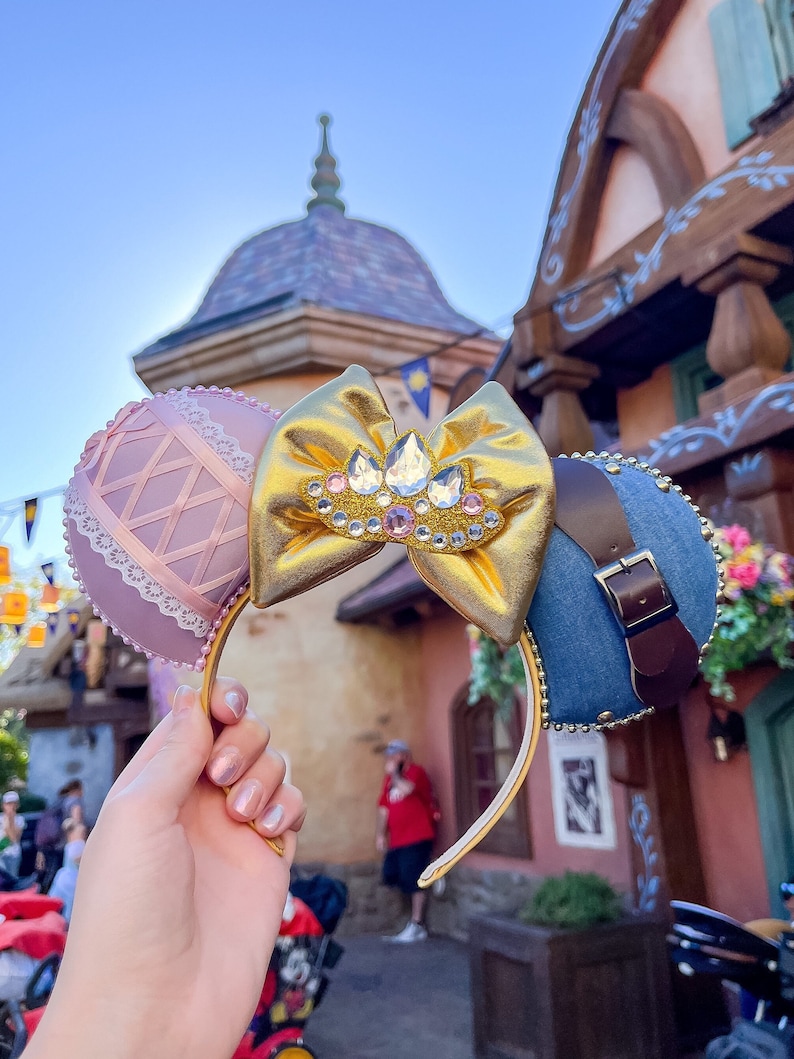 Puede incluir: Un par de orejas de Minnie Mouse rosa y azul con un lazo dorado y una corona de diamantes de imitaci&oacute;n. Las orejas est&aacute;n decoradas con un patr&oacute;n de cuadros rosa y blanco y una tela vaquera con una correa de cuero marr&oacute;n.
