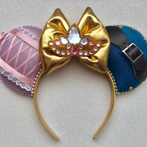 Puede incluir: Diadema dorada con orejas de tela rosa y azul. La oreja rosa tiene un borde de encaje y la oreja azul tiene una correa de cuero marr&oacute;n con una hebilla plateada. La diadema tiene un lazo dorado con una corona de diamantes de imitaci&oacute;n en el centro.