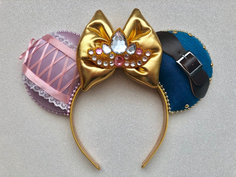 Puede incluir: Diadema dorada con orejas de tela rosa y azul. La oreja rosa tiene un borde de encaje y la oreja azul tiene una correa de cuero marr&oacute;n con una hebilla plateada. La diadema tiene un lazo dorado con una corona de diamantes de imitaci&oacute;n en el centro.