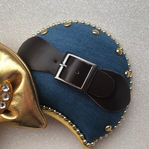 Puede incluir: Un par de orejas de rat&oacute;n de mezclilla azul con un cintur&oacute;n de cuero marr&oacute;n y una hebilla plateada. Las orejas est&aacute;n decoradas con cuentas y tachuelas doradas.