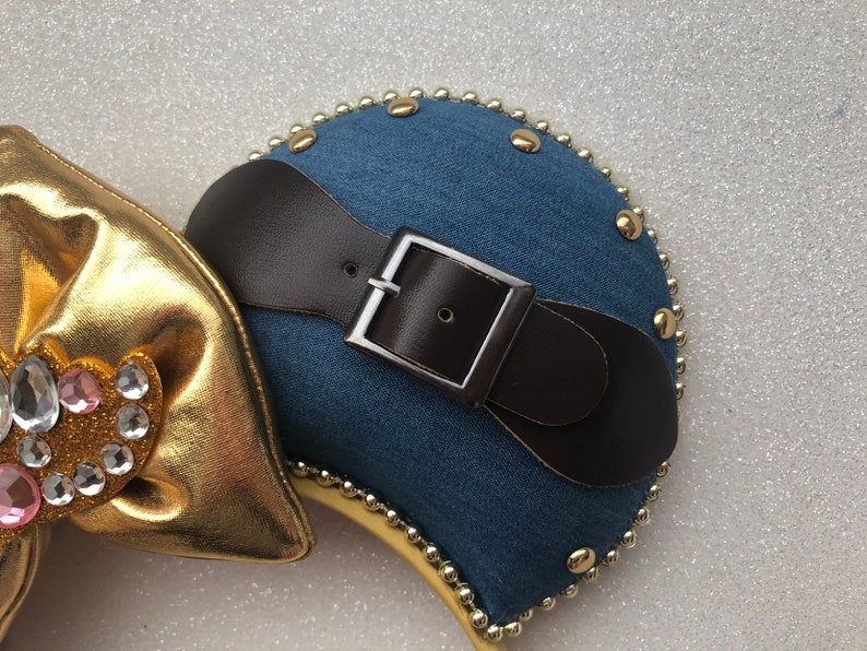 Puede incluir: Un par de orejas de rat&oacute;n de mezclilla azul con un cintur&oacute;n de cuero marr&oacute;n y una hebilla plateada. Las orejas est&aacute;n decoradas con cuentas y tachuelas doradas.