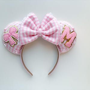 May include: A pink and white gingham-patterned headband with a large bow. The ears are decorated with a pink bow and the letter "M" in pink and gold. The headband is trimmed with white lace and pearls.