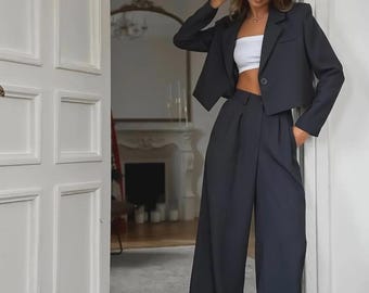 Pode incluir: Um conjunto feminino com um blazer preto curto, calças pretas de pernas largas de cintura alta e um top branco. O conjunto é usado com tênis brancos. O fundo mostra uma porta e uma sala com lareira.