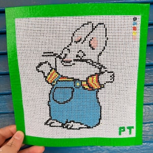 May include: Handmade needlepoint featuring a cartoon rabbit character in blue overalls and a striped shirt. The artwork is set against a white background with a green border. The initials "PT" are in the lower right corner.