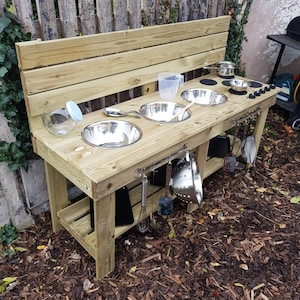 May include: A wooden outdoor play kitchen with a bench back, featuring stainless steel bowls and cooking utensils. The play kitchen includes a clear plastic measuring jug, a glass jar, and cooking tools hanging below the counter.