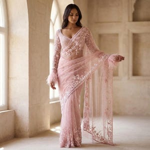 May include: A light pink saree with detailed floral embroidery and feather trim on the sleeves. The sheer fabric drapes elegantly. The saree features a detailed bodice and a matching veil. The design is suitable for special occasions.