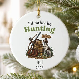 May include: A round white ceramic ornament featuring the text I'd rather be Hunting, a graphic of a rifle, a backpack, a hunting dog, a trail sign, the name Bill, and the year 2026. The ornament hangs from a gold string.