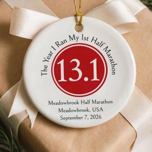 Puede incluir: Un adorno redondo de cerámica blanca con un círculo rojo y el número 13.1 en blanco. El texto dice: The Year I Ran My 1st Half Marathon, Meadowbrook Half Marathon, Meadowbrook, USA, September 7, 2026. Tiene un lazo de cuerda dorada arriba.