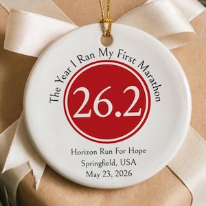 May include: A round white ceramic ornament featuring a red circle with the number 26.2 in white. The text reads The Year I Ran My First Marathon, Horizon Run For Hope, Springfield, USA, and May 23, 2026. It includes a gold string for hanging.