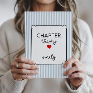 May include: A hardcover book with a light blue and white vertical striped cover. The center features a white rectangular label with a black border containing the text "CHAPTER thirty" above a small red heart and the name "emily" in black script.