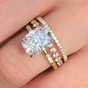 May include: A three-piece gold ring set. The engagement ring features a large, oval-cut diamond. Two additional bands, one with small diamonds and another with larger stones, complete the set. The rings are displayed on a person's finger.