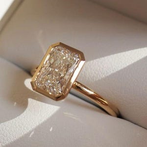 May include: A gold ring featuring a rectangular-cut diamond with a gold bezel setting. The diamond is set in a gold frame, giving a modern and elegant appearance. The ring is displayed in a white jewellery box.