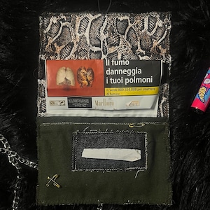 Puede incluir: Una funda de tabaco hecha a mano con solapa con estampado de piel de serpiente y cuerpo verde oscuro. La funda contiene un paquete de cigarrillos Marlboro y una etiqueta de advertencia en italiano. Se adjunta un pequeño alfiler dorado y una cadena plateada.