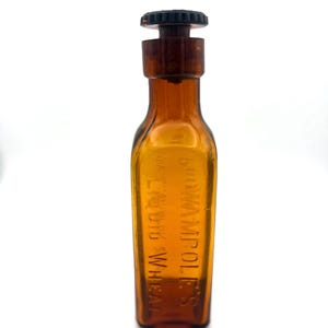 May include: An amber-colored glass bottle with a dark brown cap. The bottle has a rectangular shape with rounded shoulders and a narrow neck. The text "LIQUID WHEAT" and "BROMAMPOL'S" is printed vertically on the bottle.