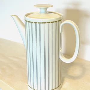 Può includere: Una caffettiera in ceramica bianca con coperchio e manico. La caffettiera presenta linee verticali nere e dettagli dorati. Il beccuccio è sul lato sinistro. Il manico è curvo.