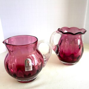 Puede incluir: Dos jarras de cristal color arándano con asas transparentes. Una jarra tiene un borde con volantes y la otra un borde liso. Ambas tienen una etiqueta que dice "Pilgrim Glass Handcrafted".
