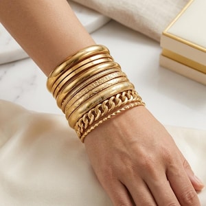 Puede incluir: Un conjunto de pulseras doradas que incluye varios brazaletes lisos, dos brazaletes con patrones geométricos grabados, una pulsera de cadena gruesa y una pulsera fina de cuerda trenzada, todas usadas juntas en la muñeca.