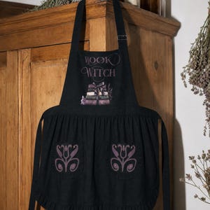 Peut inclure: Un tablier noir portant l'inscription "BOOK WITCH" au-dessus d'une illustration d'une pile de livres attach&eacute;s par un ruban. Le tablier comporte deux poches avant, chacune orn&eacute;e d'un motif floral violet sym&eacute;trique, et un ourlet &agrave; volants.