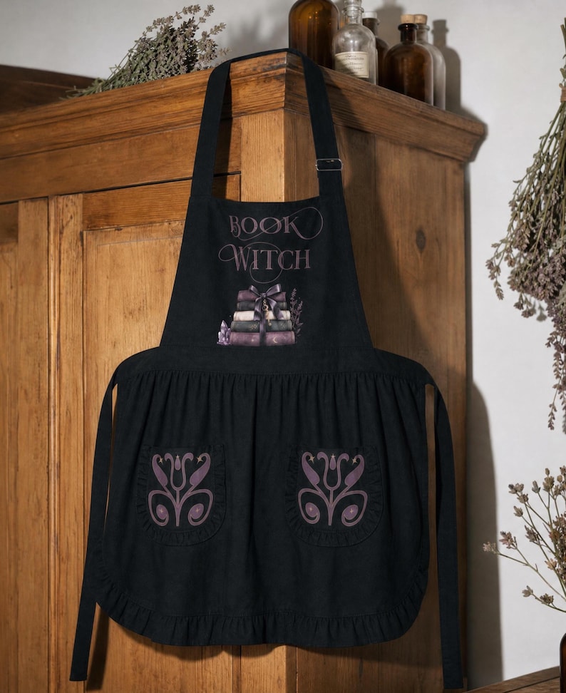 Peut inclure: Un tablier noir portant l'inscription "BOOK WITCH" au-dessus d'une illustration d'une pile de livres attach&eacute;s par un ruban. Le tablier comporte deux poches avant, chacune orn&eacute;e d'un motif floral violet sym&eacute;trique, et un ourlet &agrave; volants.