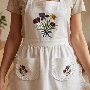 May include: White apron with a floral bouquet design. The apron features a large central bouquet of purple irises, red poppies, and yellow daffodils tied with a blue ribbon. Two pockets with floral embroidery.