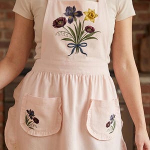 May include: A pale pink apron with two front pockets and floral embroidery. The embroidery shows purple irises, yellow daffodils, and burgundy flowers tied with a blue ribbon. The apron is made of a soft material.