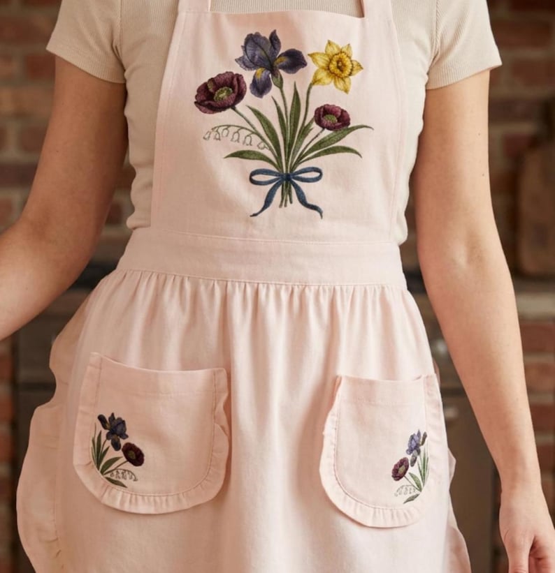 May include: A pale pink apron with two front pockets and floral embroidery. The embroidery shows purple irises, yellow daffodils, and burgundy flowers tied with a blue ribbon. The apron is made of a soft material.