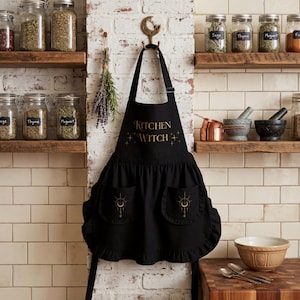 Op de afbeelding: Een zwart schort met de gouden tekst Kitchen Witch op de borst en twee geplooide zakken met gouden maan- en stergraphics. Het schort heeft een nekband en taillebanden en hangt aan een gouden muurhaak in de vorm van een halve maan.