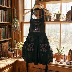Peut inclure: Un tablier noir portant l'inscription "BOOK WITCH" au-dessus d'un motif de livres empil&eacute;s. Le tablier comporte deux poches avant, chacune orn&eacute;e d'un motif floral violet, et un ourlet &agrave; volants sur le bord inf&eacute;rieur de la jupe.