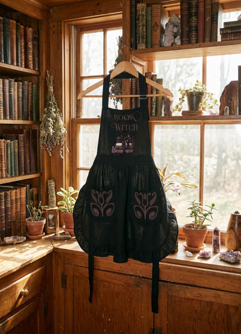 Peut inclure: Un tablier noir portant l'inscription "BOOK WITCH" au-dessus d'un motif de livres empil&eacute;s. Le tablier comporte deux poches avant, chacune orn&eacute;e d'un motif floral violet, et un ourlet &agrave; volants sur le bord inf&eacute;rieur de la jupe.