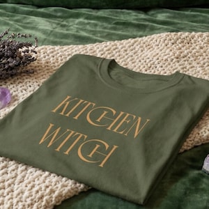 May include: An olive green t-shirt featuring the text KITCHEN WITCH printed in a gold-colored serif font across the center of the chest. The garment has a classic crew neckline and short sleeves, presented in a flat, folded orientation.