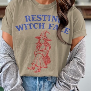 Könnte beinhalten: Beiges T-Shirt mit dem blauen Schriftzug "RESTING WITCH FACE". Darunter eine rote Illustration einer Hexe mit Hut. Das T-Shirt wird mit einer blauen Jeans und einer grauen Strickjacke getragen.