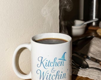 Peut inclure: Une tasse en céramique blanche remplie de café noir, de la vapeur s'élevant du dessus. La tasse porte l'inscription "Kitchen & Witchin'" en script bleu clair, ainsi qu'une illustration de chapeau de sorcière. La tasse est posée sur une surface en bois.