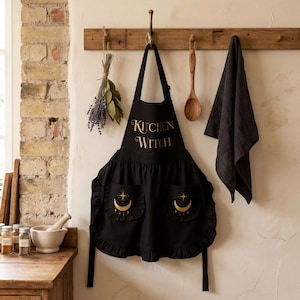 May include: Black apron with the text "Kitchen Witch" in gold script. The apron has two pockets with a crescent moon and star design. A wooden spoon and a black towel hang nearby.