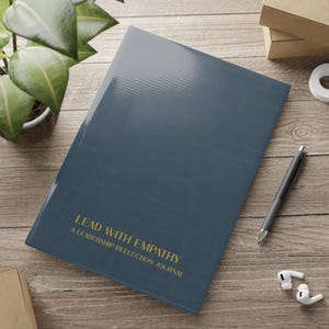 Könnte beinhalten: Ein marineblaues Notizbuch mit dem goldenen Text "Lead with Empathy: A Leadership Reflection Journal" liegt auf einer Holzoberfläche. Ein schwarzer Stift und weiße kabellose Ohrhörer liegen neben dem Notizbuch. Eine Topfpflanze ist im Hintergrund.