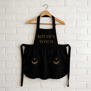 May include: Black apron with the words "KITCHEN WITCH" in gold script. The apron has two pockets with a crescent moon and star design. The apron is hanging on a wooden hanger against a white brick wall.
