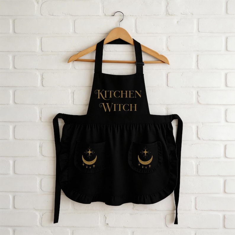 May include: Black apron with the words "KITCHEN WITCH" in gold script. The apron has two pockets with a crescent moon and star design. The apron is hanging on a wooden hanger against a white brick wall.