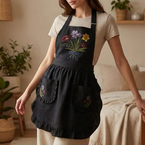 May include: Black apron featuring a floral bouquet embroidery on the bib and pockets. The apron has a ruffled hem and pockets. The embroidered bouquet includes purple, red, and yellow flowers tied with a blue ribbon. The apron is made of a dark fabric.